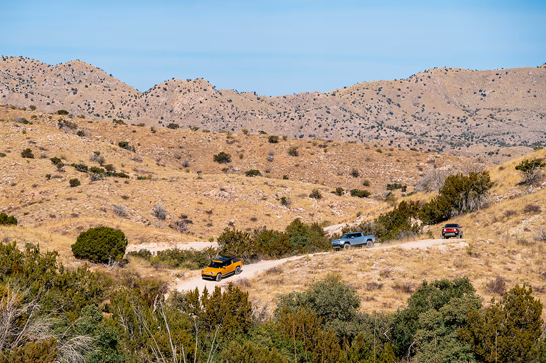 Three off-road vehicles driving on a winding dirt trail through a dry, mountainous landscape with sparse bushes under a clear blue sky.